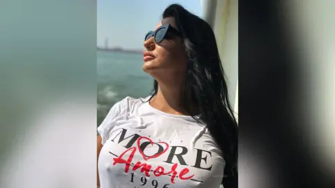 MissyJolie poses outdoors in a white graphic t-shirt and black sunglasses with her long black hair down.
