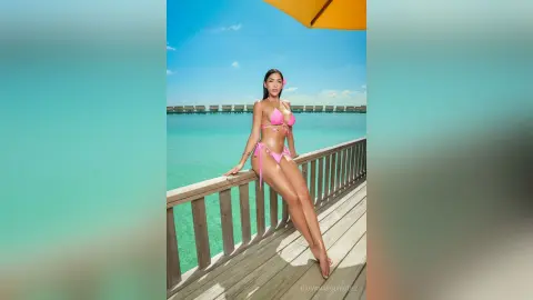 Glamorous woman in a pink bikini posing on a tropical wooden pier overlooking turquoise ocean waters.