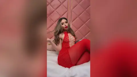Glamorous woman in a red sequin bodysuit and tights posing on a white bed in a pink room.