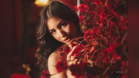 Young woman with wavy hair posing amidst vibrant red flowers in a warm, moody indoor setting.