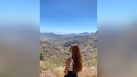 Woman in white tank top gazing at scenic mountain valley landscape under a clear blue sky.