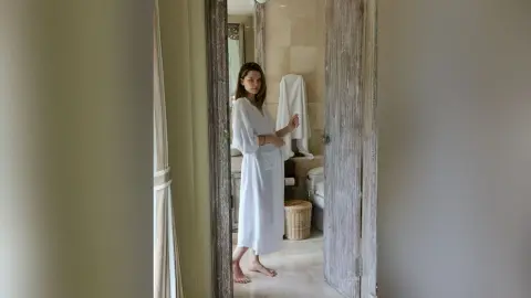 Young woman in a white spa robe standing in a bright, modern bathroom doorway.