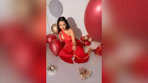 Glamorous woman in a red jumpsuit sitting among red and gold balloons in a festive celebratory setting.