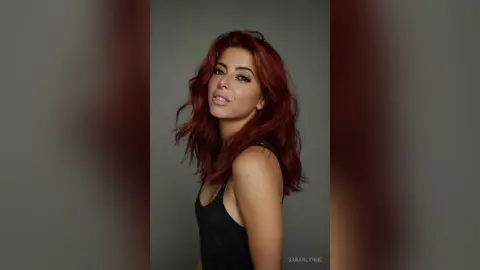 Redheaded woman in a black tank top posing with a confident expression against a grey studio background.