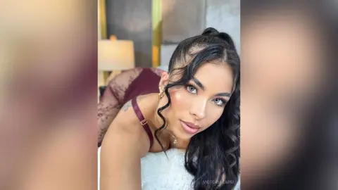 VictoriaCapri poses with long dark wavy hair, wearing a white lace top and a maroon lace bodysuit or bra strap, looking directly at the camera.