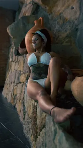 Smiling woman in white bikini sitting on a rustic stone wall in a warm, Mediterranean setting.