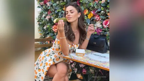 AnaMathers wears an orange and white citrus-print sundress while sitting at a table against a wall of colorful flowers.