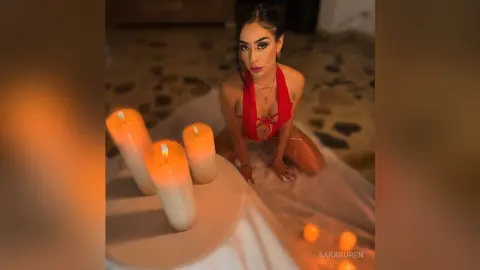 Glamorous woman in red bikini sitting by glowing candles in a warm, moody indoor setting.