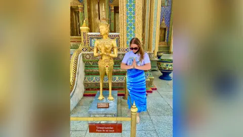 Woman in blue skirt praying beside a golden Buddha statue at a serene Thai temple.