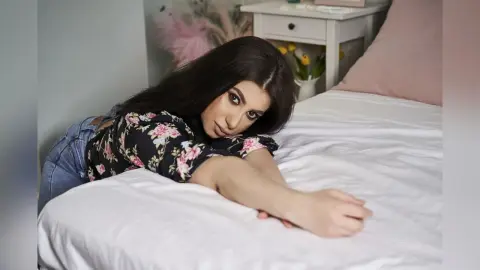 Young woman in floral blouse relaxing on a white bed in a bright, cozy bedroom setting.