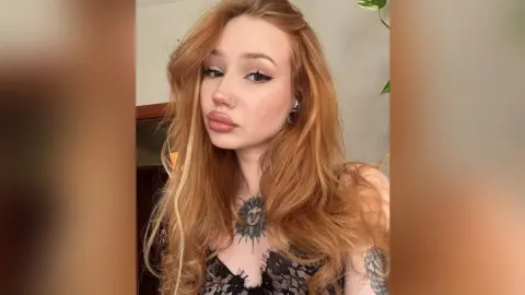 Redheaded woman with long wavy hair and chest tattoos wearing a black lace top in a soft indoor setting.