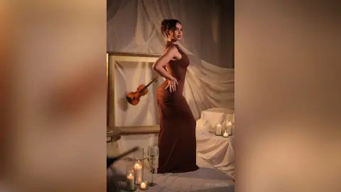 Elegant woman in a brown evening gown posing in a moody, candlelit room with a vintage violin.