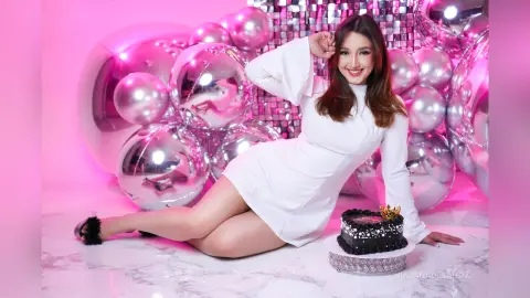 JulianaDeLaHoz smiles while sitting on a white floor next to a black cake, wearing a white long-sleeved mini dress against a background of silver disco balls.