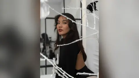 Young woman in a black outfit posing amidst white decorative branches in a minimalist modern studio setting.