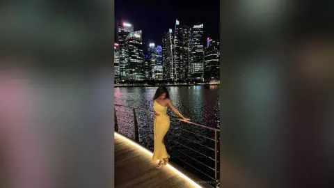 Elegant woman in a yellow evening gown standing on a wooden pier overlooking a glowing city skyline at night.