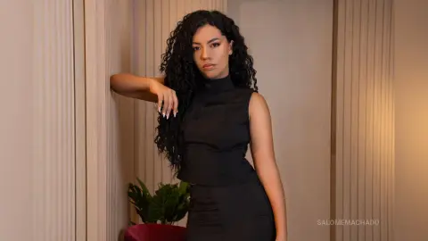 Elegant woman with long curly hair in a black sleeveless dress leaning against a neutral wall in soft lighting.