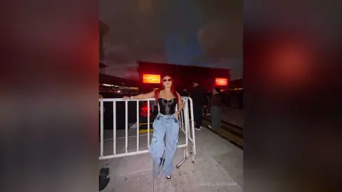 Stylish woman in a black corset and baggy jeans posing at an outdoor night event near a metal barrier.