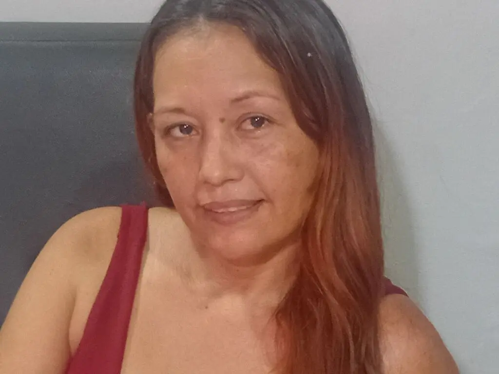 Smiling woman with long brown hair wearing a red tank top in a relaxed indoor setting.