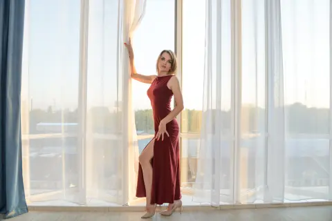Elegant woman in a long burgundy evening gown standing by bright sheer curtains in a sunlit modern room.