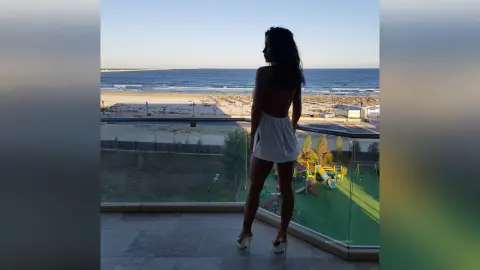 Woman in white summer outfit standing on a balcony overlooking a sunny beach and ocean view.