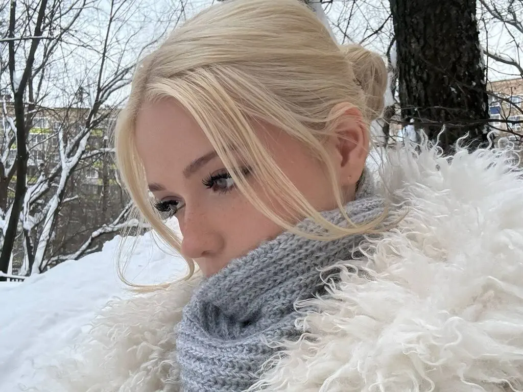 Blonde woman in a cozy white faux fur coat and grey knit scarf posing in a snowy winter forest setting.