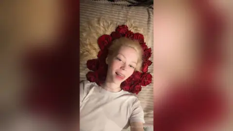Smiling blonde woman in a white t-shirt lying on a bed surrounded by red rose petals for a romantic mood.