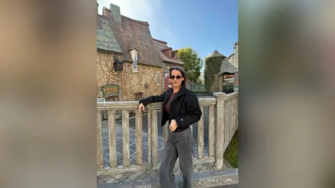 Stylish woman in a black jacket and grey jeans leaning on a wooden fence in a quaint European village.