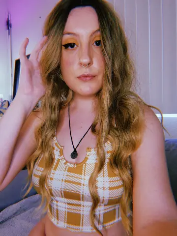 Young woman with long wavy hair wearing a yellow plaid crop top and necklace in a cozy indoor setting.