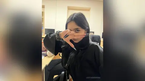 Young woman in a black long-sleeve outfit making a peace sign in a modern indoor setting.