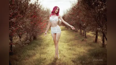 Young woman with pink hair in white lace lingerie standing in a dreamy, sunlit meadow surrounded by trees.