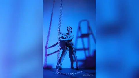 Fashionable woman in edgy black bodysuit posing with metal chains in moody blue studio lighting.