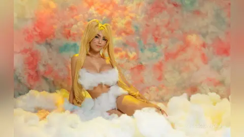 Blonde woman in a white fluffy cloud outfit sitting against a dreamy pastel colorful cloud background.