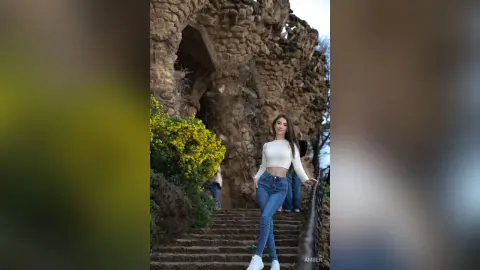 Young woman in white crop top and blue jeans standing by stone ruins in a sunny garden setting.