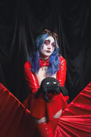 KimLennoxc wears a red latex outfit and blue hair while posing with a large black spider plushie against a black curtain.