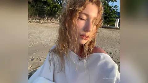 Blonde woman in a white linen shirt relaxing on a sunny beach at golden hour.