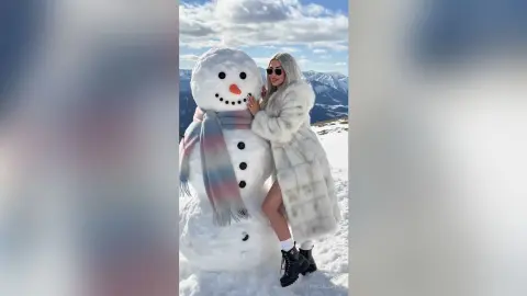 Stylish woman in a white faux fur coat posing with a large snowman in a sunny snowy mountain landscape.