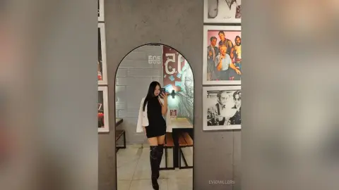 EvieMillers poses in a black dress and long black boots, standing in front of a mirror in a room with grey brick walls and framed photos.