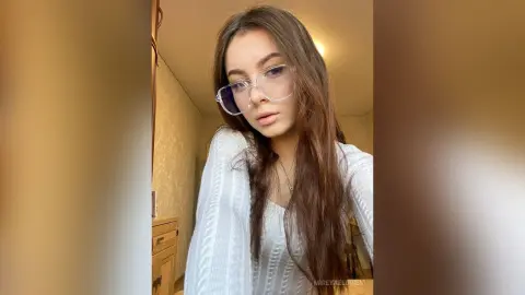 Young woman with long brown hair wearing clear glasses and a white ribbed sweater in a warm indoor setting.