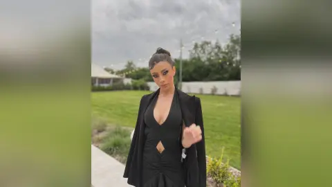 Elegant woman in a black cutout evening gown and blazer standing in a lush green garden under a cloudy sky.