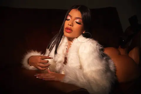 Glamorous woman in a white faux fur coat relaxing on a dark sofa with elegant makeup and hoop earrings.