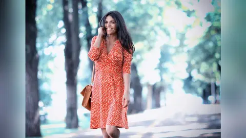 Smiling woman in a red polka dot summer dress walking through a sunny, leafy park.