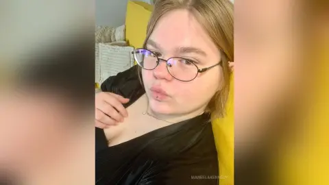 ManeelaKennedy wearing glasses and a black top, posing with a pouty expression while leaning back against a yellow pillow.