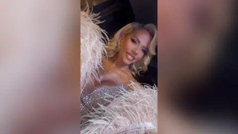 Smiling blonde woman in a glamorous silver sequin gown with white feather trim for a luxury gala event.
