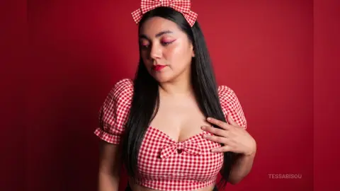 Young woman in red gingham crop top and matching bow headband posing against a red background.