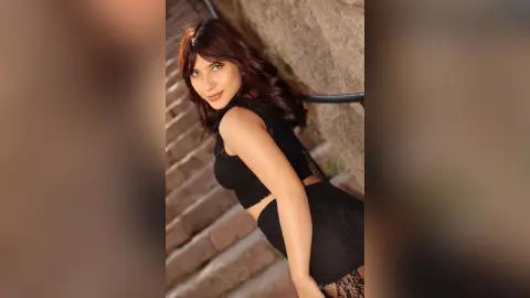 Smiling young woman in a black lace outfit sitting on stone stairs in warm sunlight.