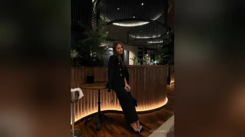 EvelynMills stands in a dark, modern lounge wearing a black button-up blazer and matching trousers.