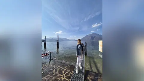 Stylish woman in a grey hoodie and white pants standing on a wooden pier by a calm lake under a blue sky.