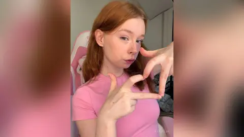 Redheaded woman in a pink t-shirt making a heart gesture in a bright gaming room setting.