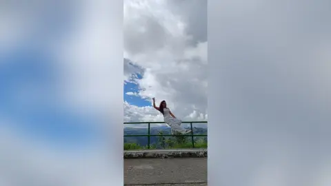 AmelieLautrec poses outdoors in a white sleeveless top and white pants, leaning back against a green metal railing under a cloudy sky.
