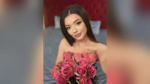 Beautiful woman with long dark hair holding a large bouquet of pink roses in a cozy bedroom setting.